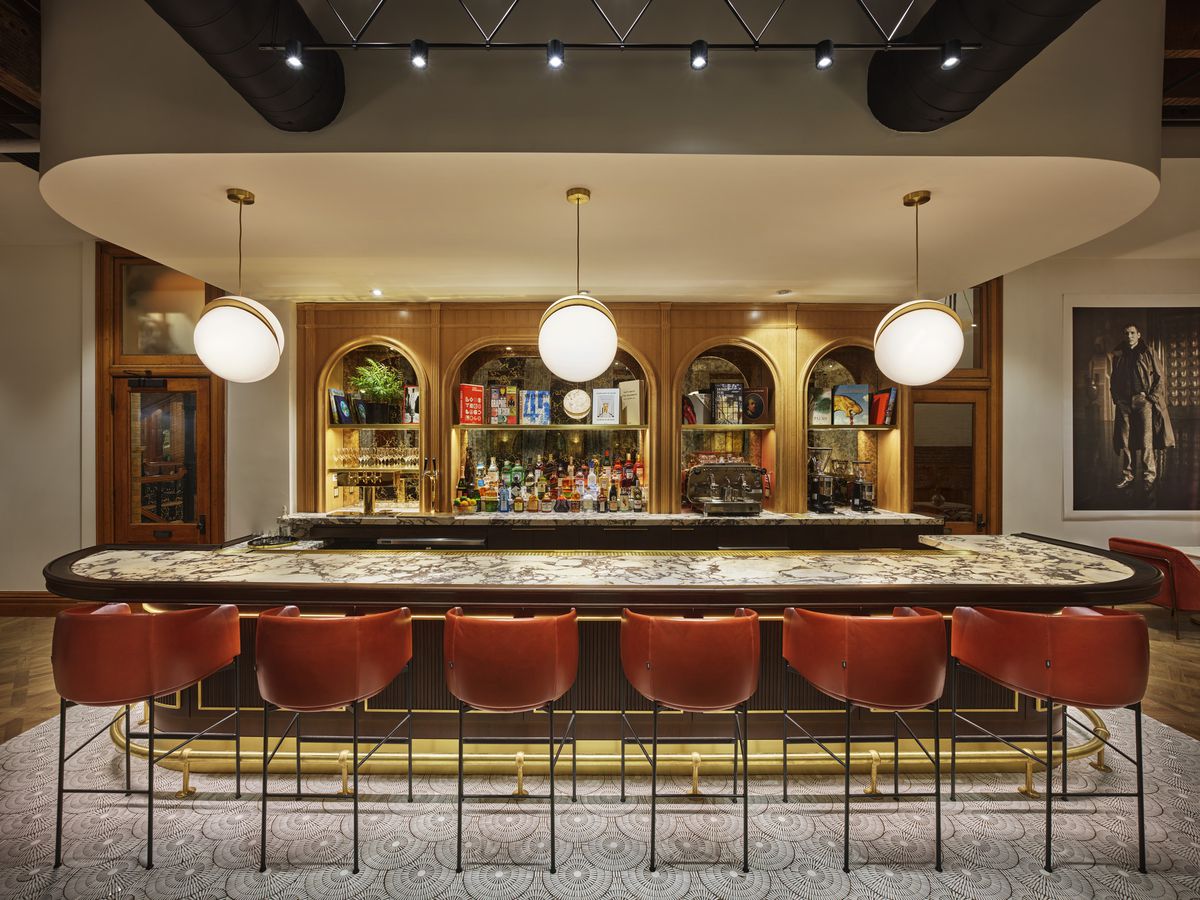 A long marble bar and tufted red bar seats inside of a dimming room with wood and brick.