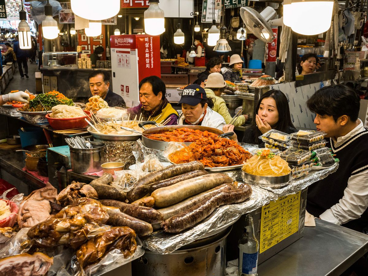 Diners sit at a stand inside a busy food market. The counter is overloaded with various dishes. 