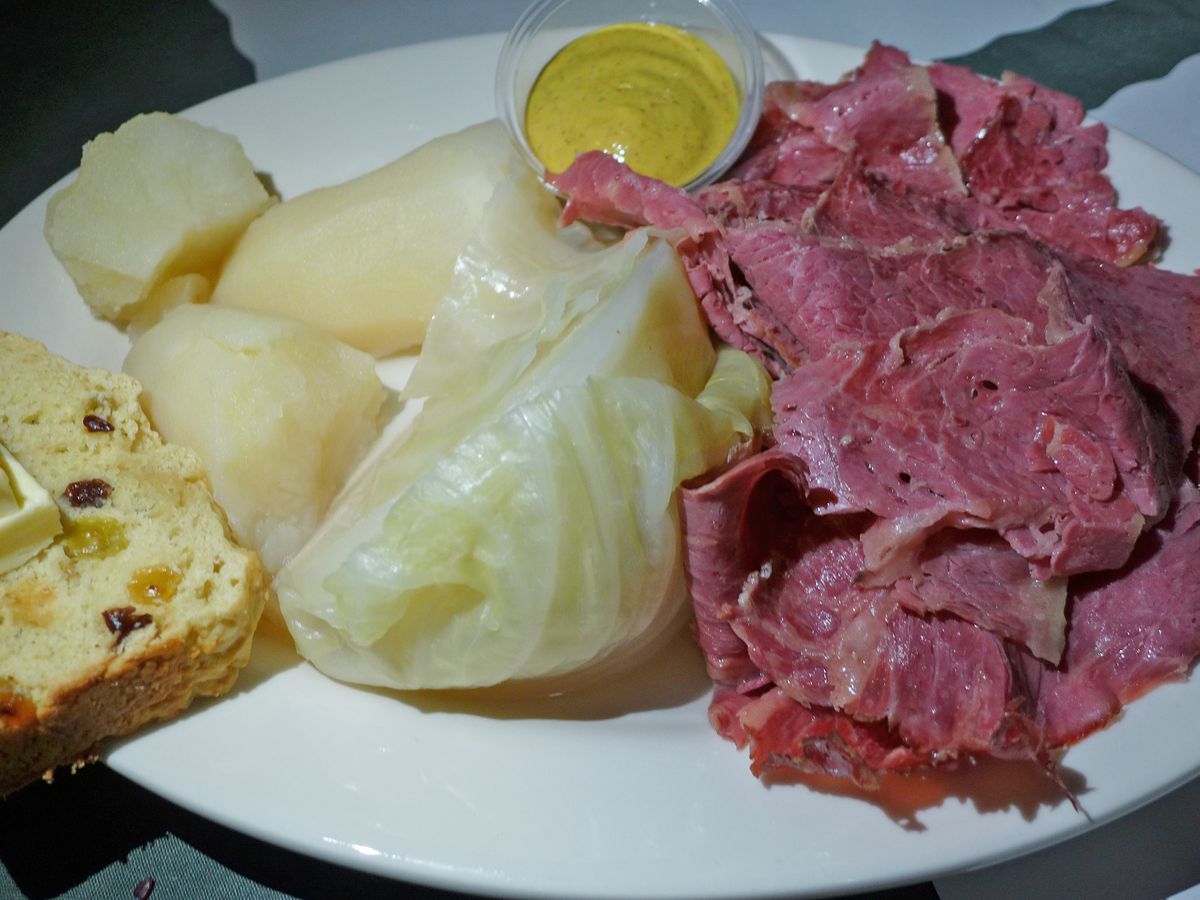An oblong bone white plate with red meat, white potatoes, and greenish cabbage, with mustard and Irish soda bread on the side.
