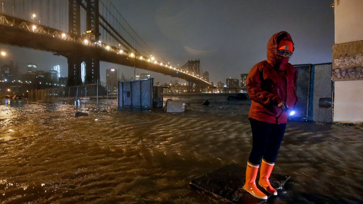 A woman stands on plywood while her surroundings are flooded with water. A bridge can be seen in the distance