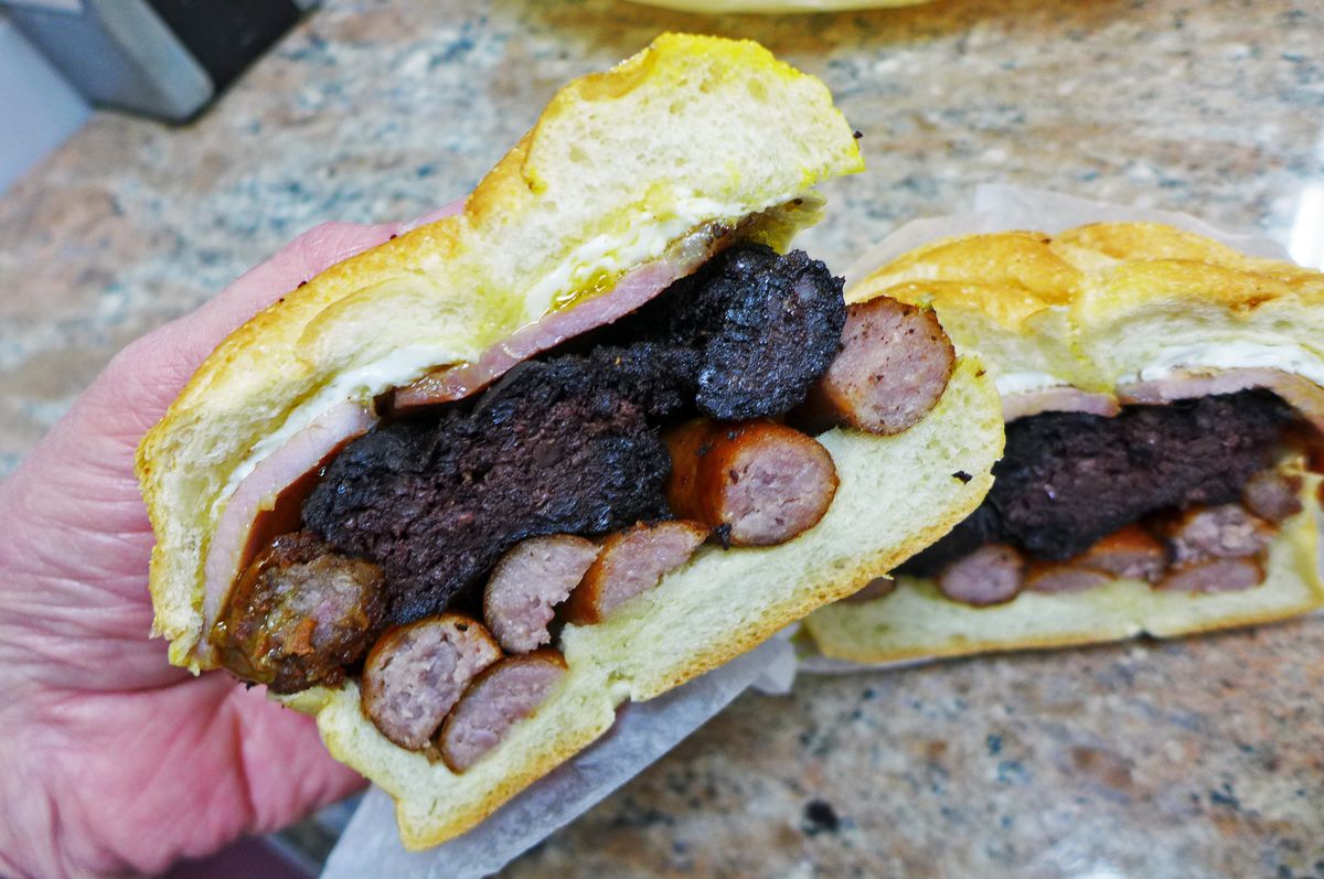 A hand holds half the sandwich, so all the components inside can be seen in their distinct layers, with blood sausage predominating.
