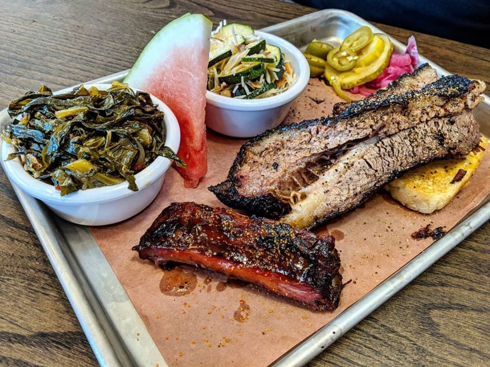 An array of barbecue items are on a piece of brown paper on a metal tray —&nbsp;brisket, a rib, collard greens, a slice of watermelon, and more
