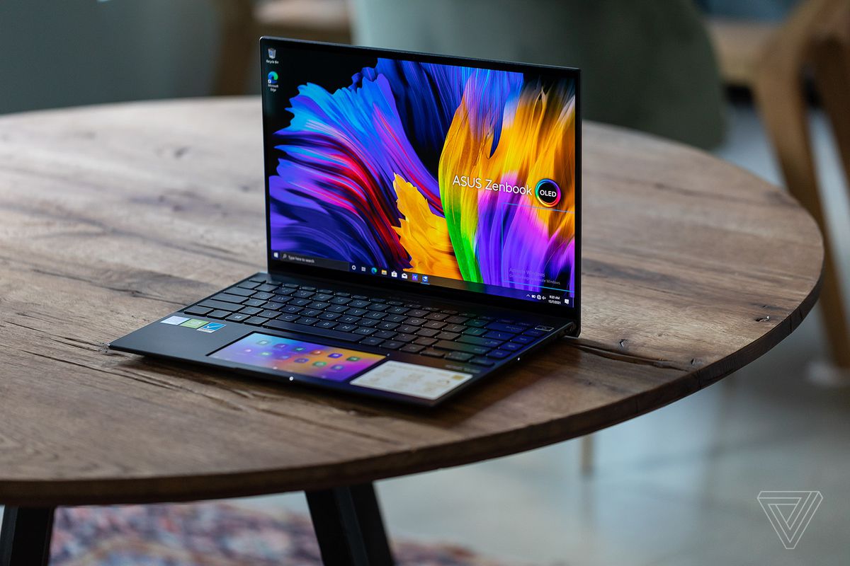 The Asus Zenbook 14X OLED on a wooden table angled to the left. The screen displays a multicolor background with the Asus Zenbook OLED logo. The touchpad displays the Screenpad homepage.