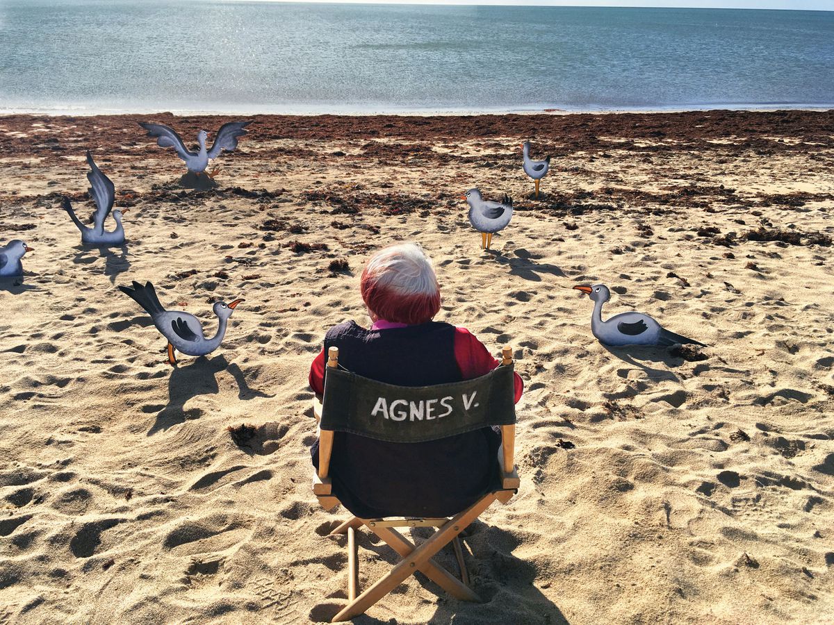 Agnès Varda sitting in a director’s chair on the beach flanked by wooden bird standups.