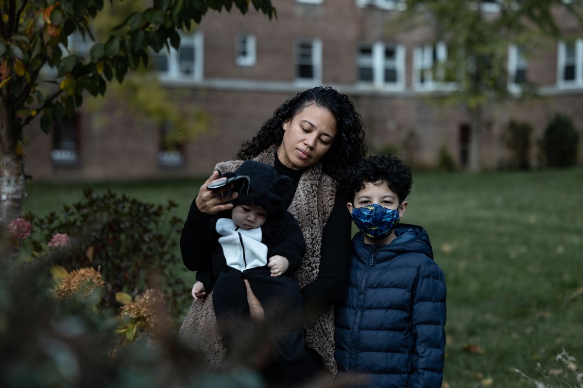 A woman with curly black hair holds her infant child, with her son standing next to her wearing a blue jacket and a protective mask. There are leaves in the foreground and a brick building behind them.