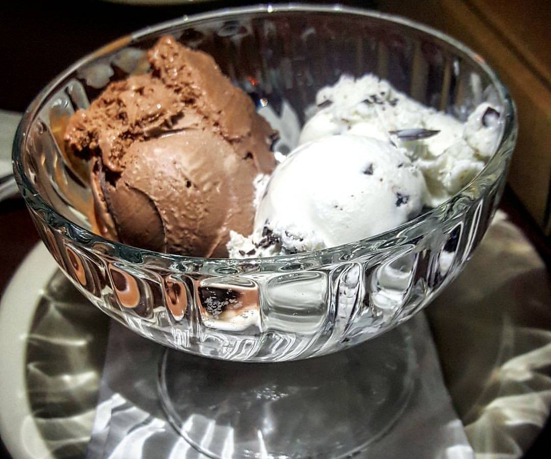 A scoop of chocolate ice cream and a scoop of mint chip ice cream sit side by side in a big glass bowl on a white plate