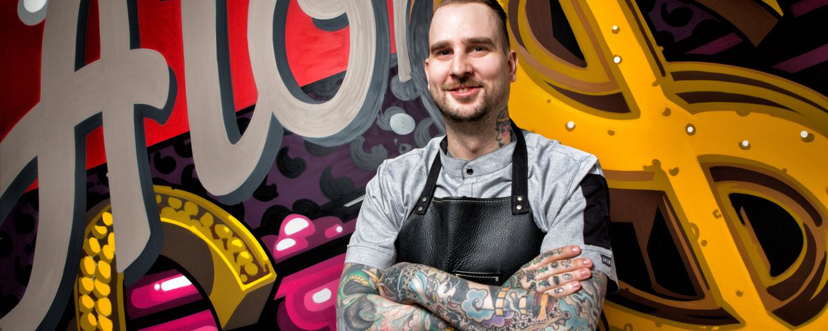 A chef with tattoos stands in front of a graffiti wall