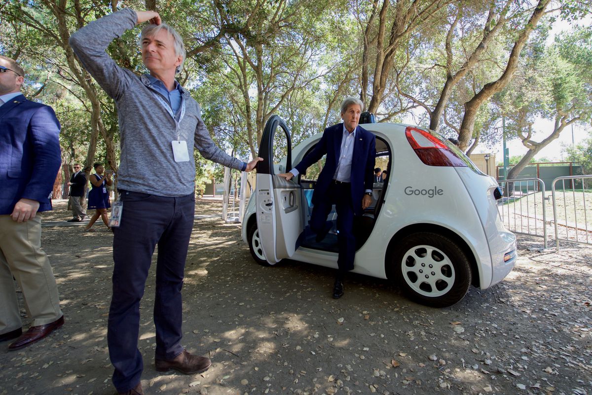 John Kerry stands with one foot out of a white Google-branded car.