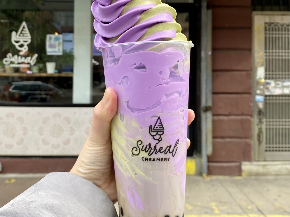 A taro bubble tea topped with a swirl of matcha-taro ice cream is held up by a hand.