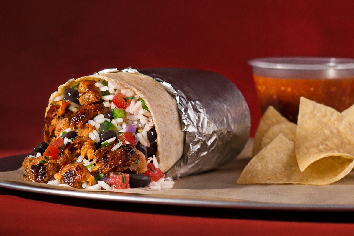 A Chipotle chorizo burrito next to a bag of chips and a plastic container of salsa, up against a red backdrop.