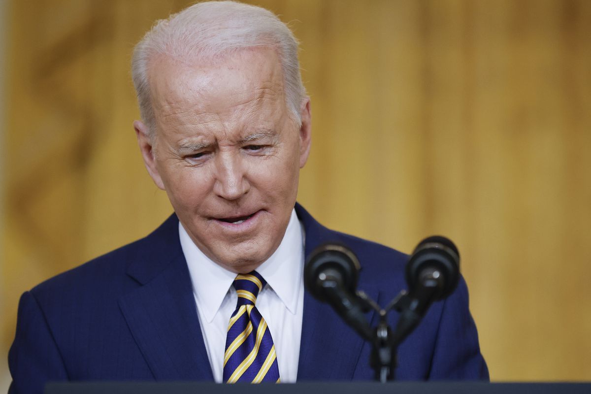 President Joe Biden speaks into a set of microphones during a press conference, wearing a dark blue suit and striped tie, in front of a gold backdrop.