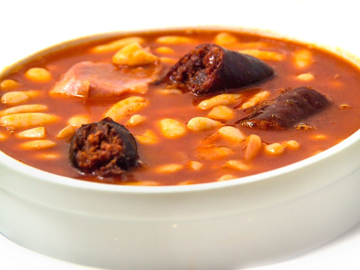 Bowl of stew with beans, sausage, and meat breaking the surface of deep red broth. 