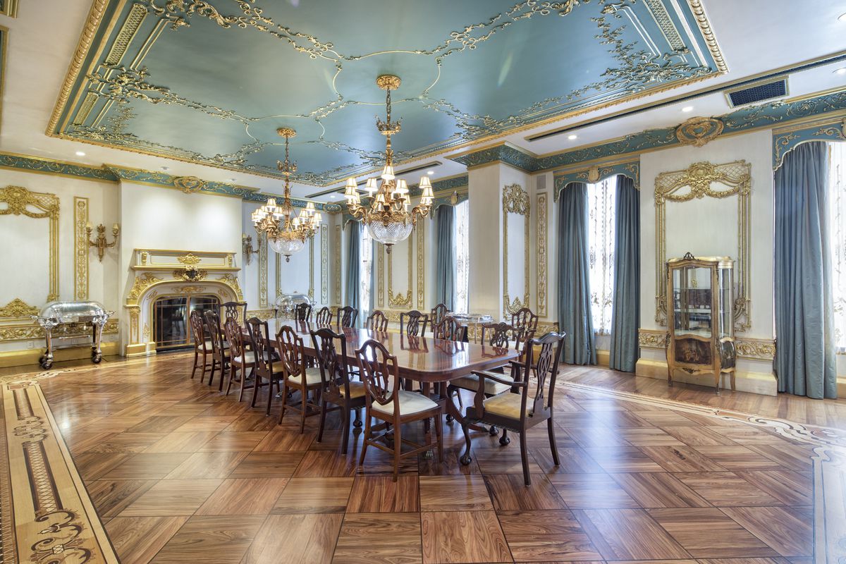 A dining room with hardwood floors, a large wooden table with chairs, large windows with blue curtains, two chandeliers, and a wood-burning fireplace.