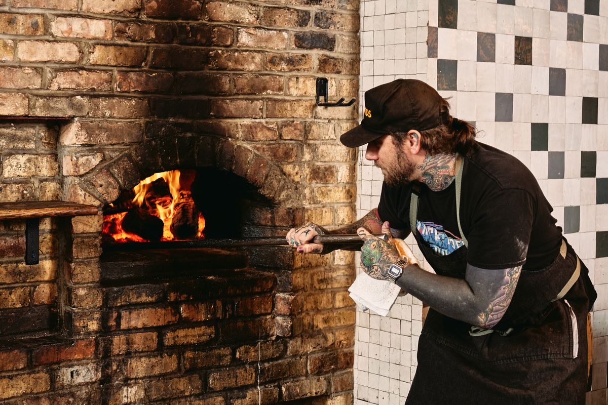 A chef with sleeve tattoos holding a paddle into a wood-fire brick oven.