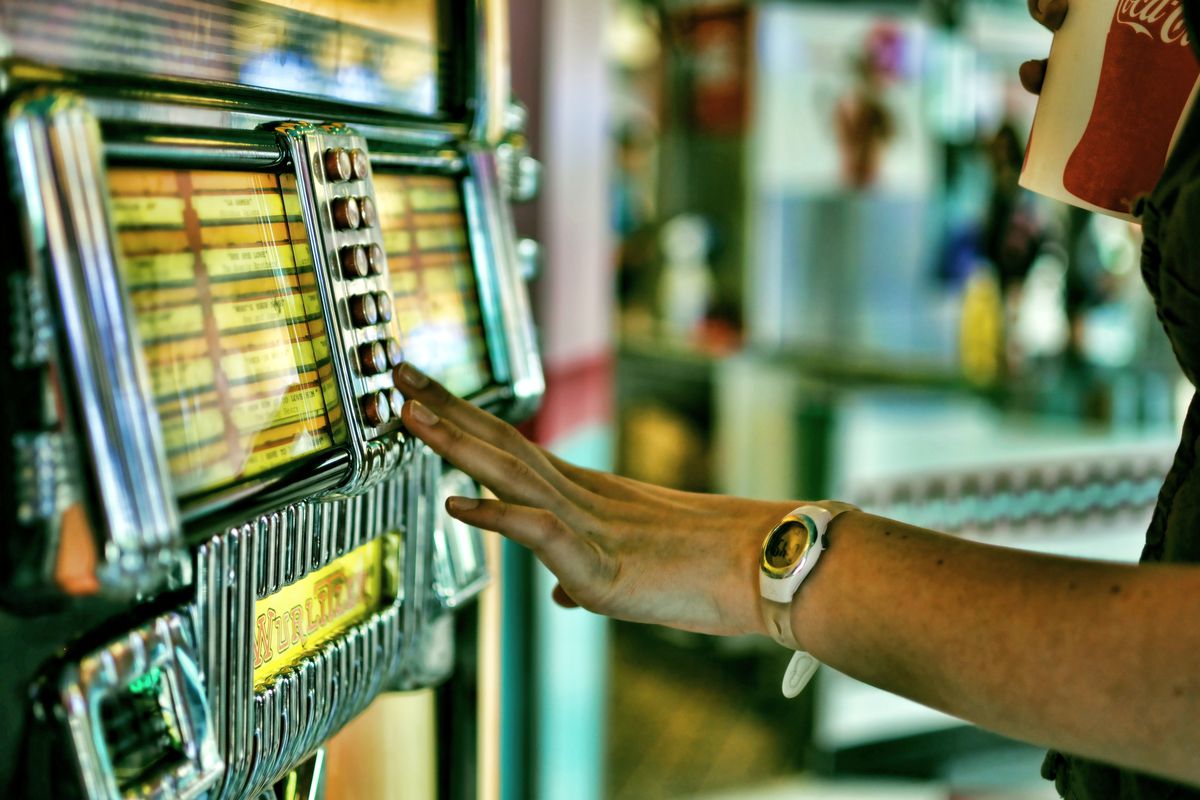 Hand reaches to touch a button on a juke box, with the other hand in the corner holding a Coke cup, with a restaurant in the background