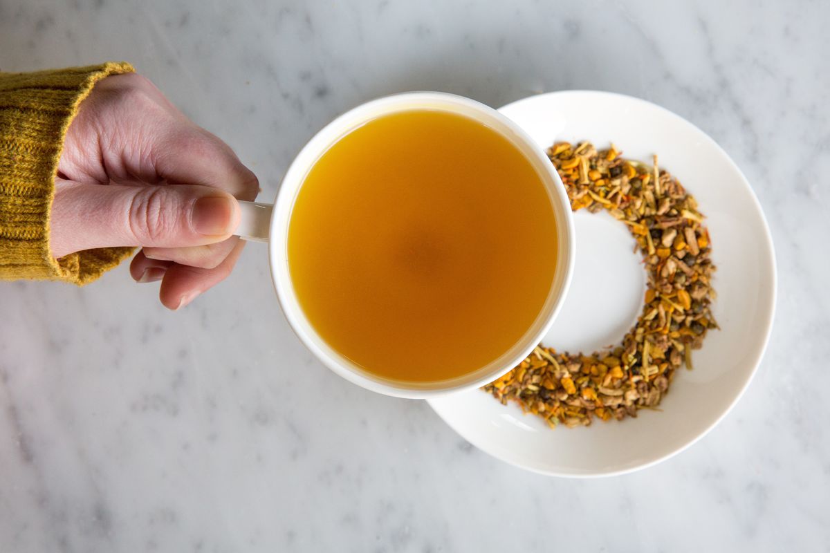 A hand holds a mug of orange-brown tea over a floral-looking pile of loose-leaf tea