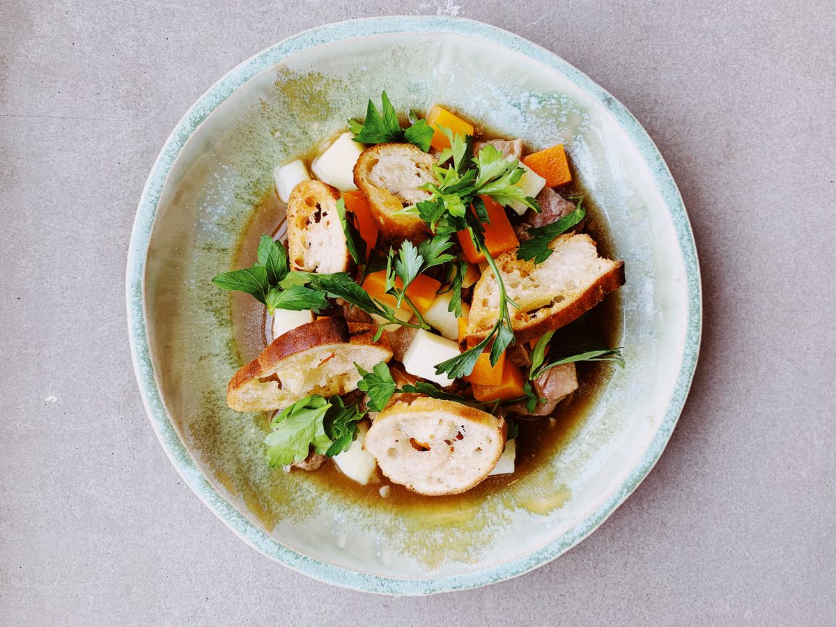 From above, a ceramic bowl on a pastel background, with slices of pork belly, carrots, parsnips, croutons, and parsley in a shallow broth