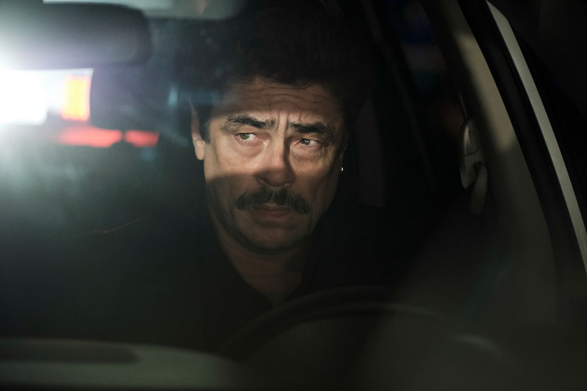 Benicio Del Toro as Tom Nichols sitting behind the wheel of a vehicle at night with headlights shining through his back window in Reptile.