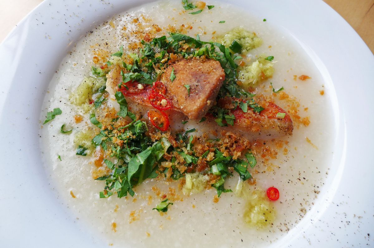 A limpid rice soup with fish, red chiles, and green herbs visible, in a white shallow bowl.