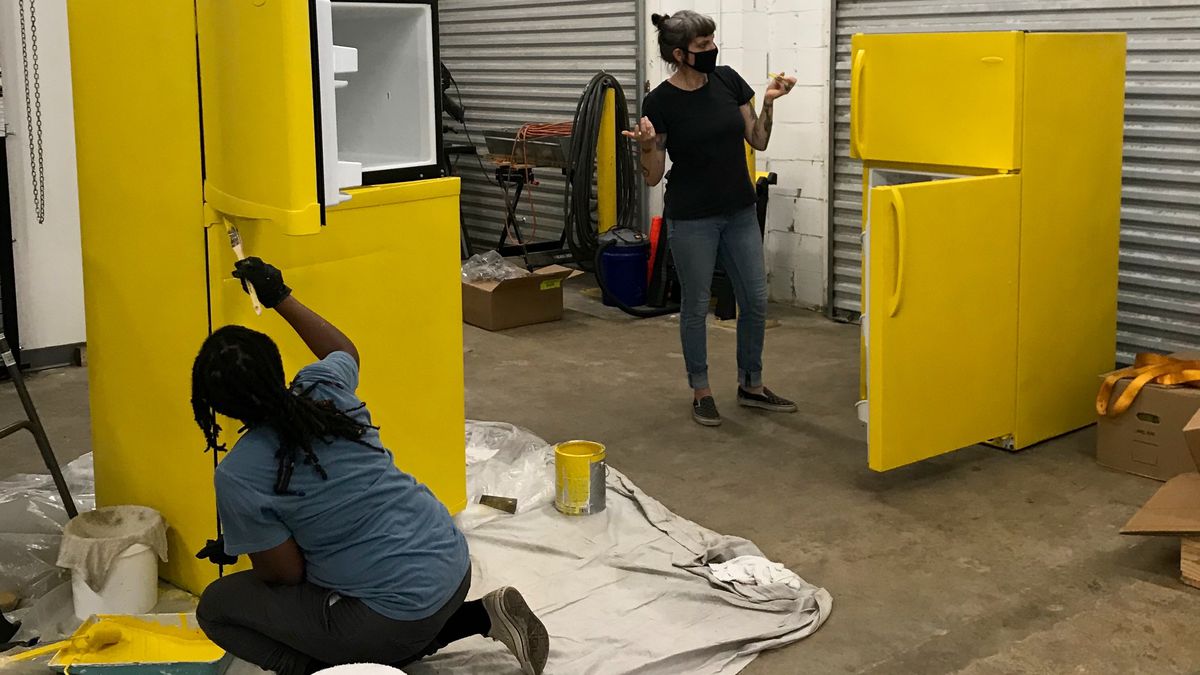 Two volunteers, one African American woman kneeling on the left and one white woman standing on the right and both in masks, painting two donated refrigerators yellow in a large white garage