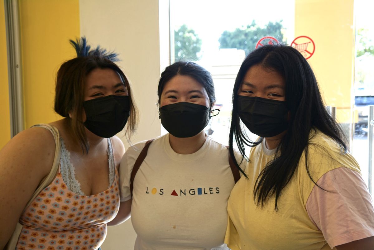 Three women in face masks smile at the camera in a boba shop.
