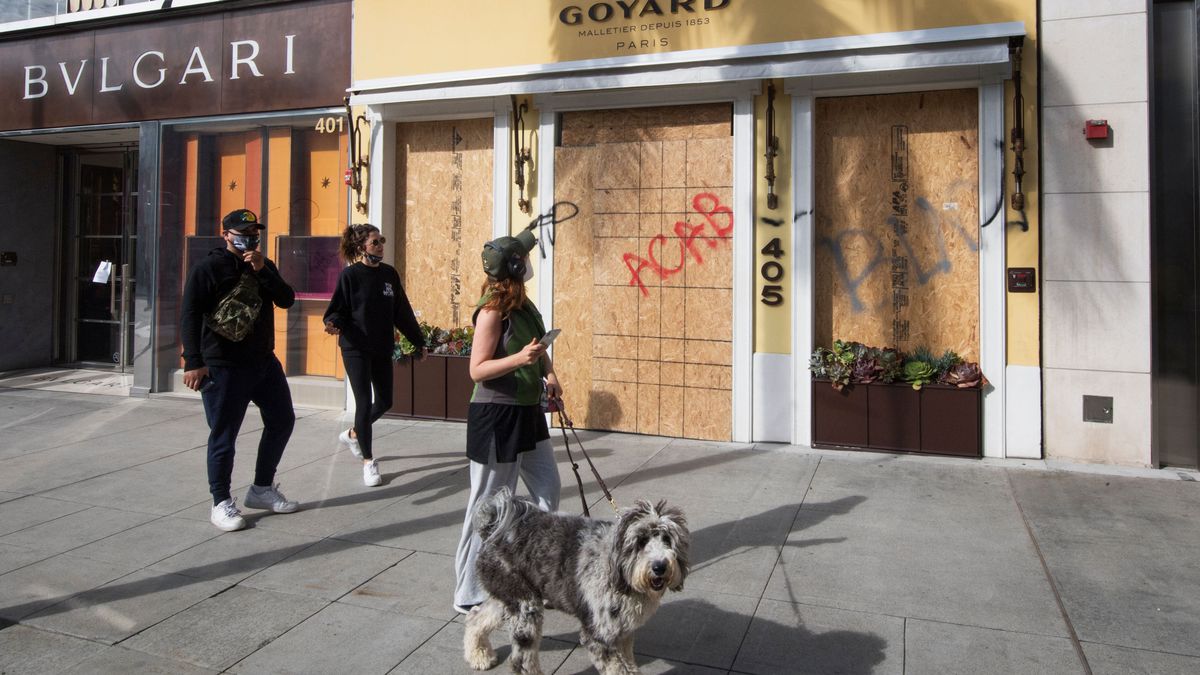 A woman with a gray white dog on a leash looks up at a boarded up Goyard storefront spray-painted with “ACAB.” 