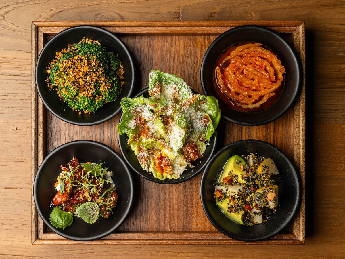 An overhead shot of five black bowls of vegetable snacks on a wooden board at LA restaurant Yangban.