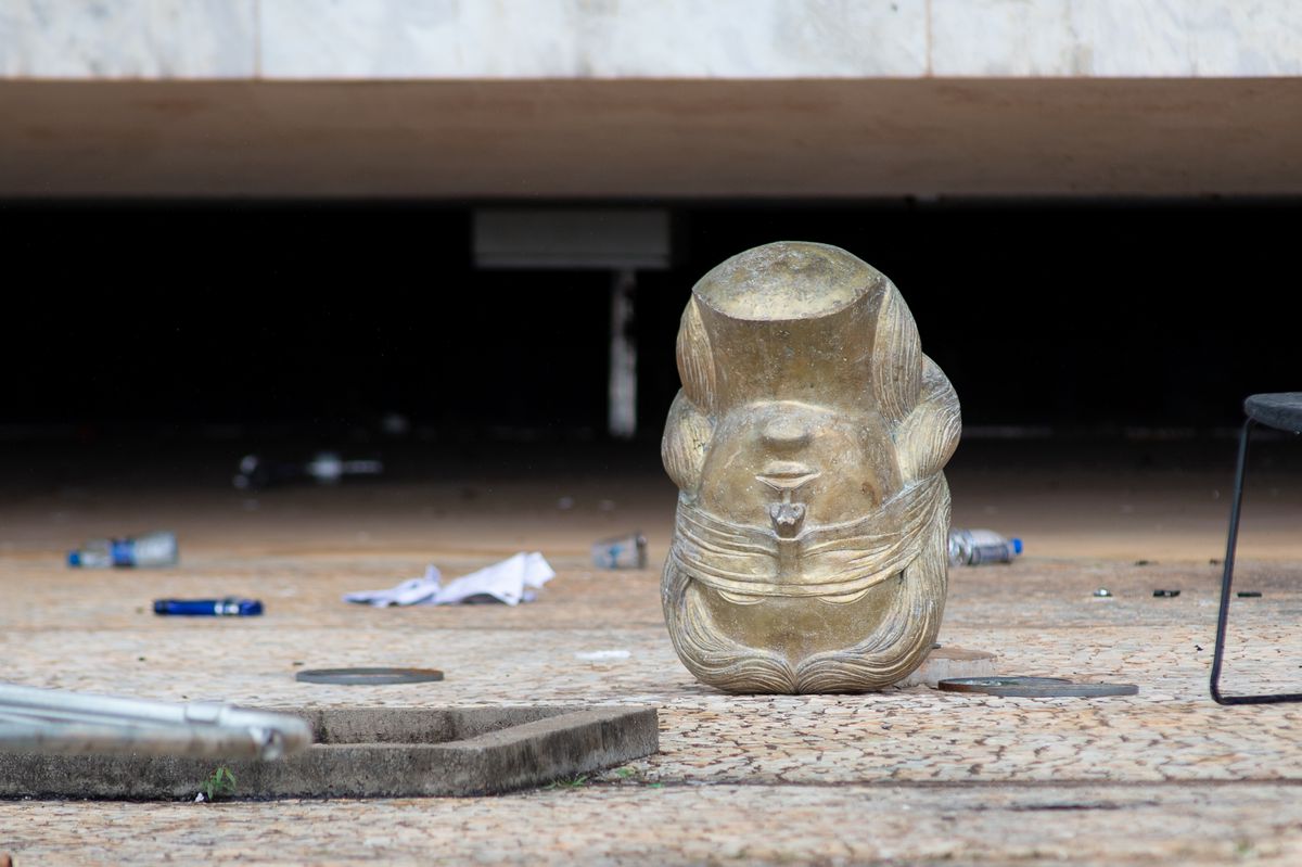 A photo showing a sculpture. The sculpture is of the head of a woman with a blindfold over her eyes. It is upside-down on a floor littered with trash.