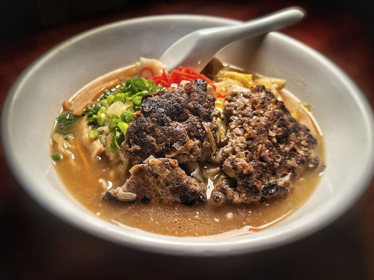 A bowl of ramen is shown against a dark wooden background. On top of the broth sits a large, broken ground beef patty.