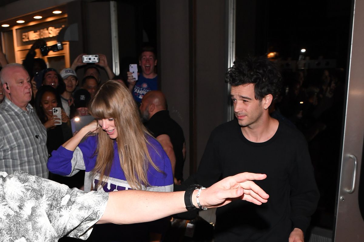 A photo shows Taylor Swift and Matty Healy exiting a glass door onto a crowded sidewalk, side by side. A person otherwise unseen is throwing an arm in front of the pair to keep onlookers away.