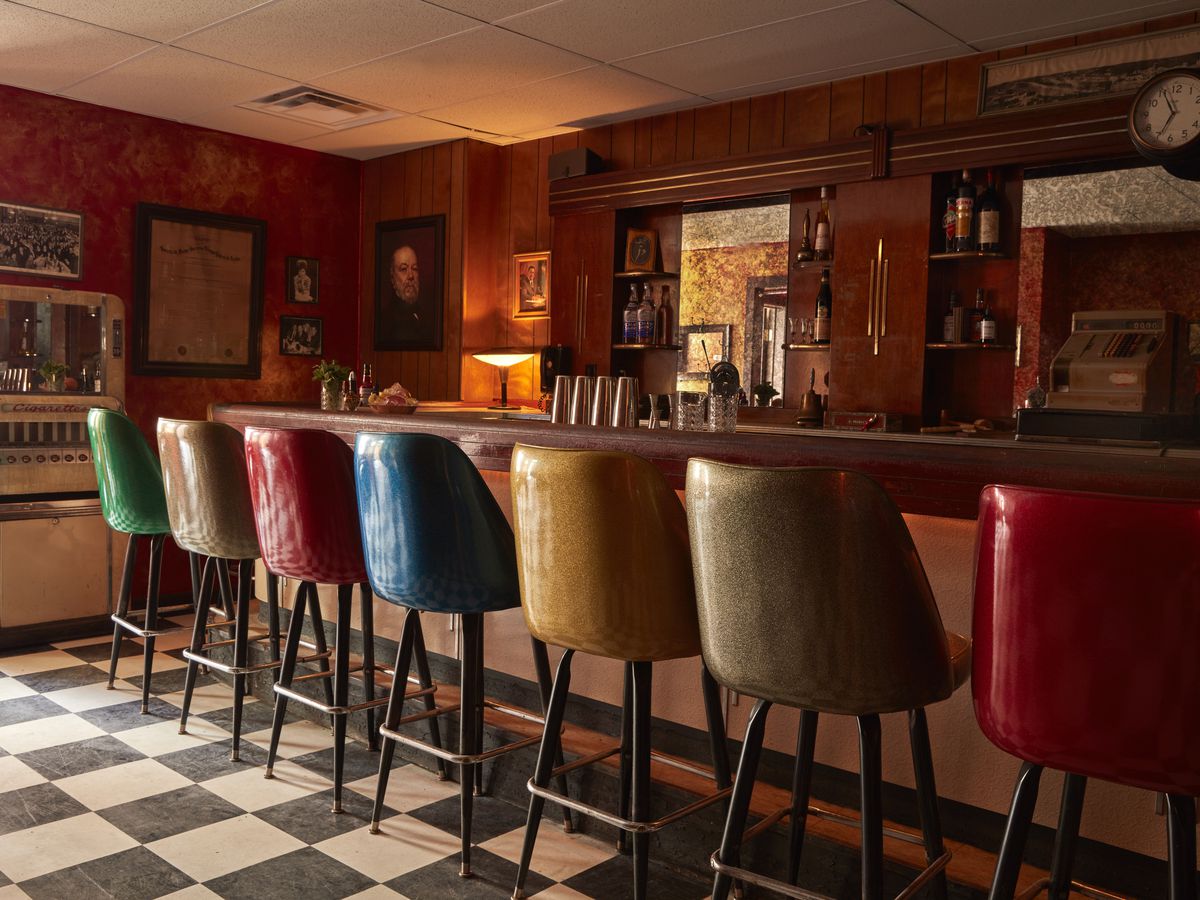The backs of stools in all different colors lined up at a bar with a white-and-black-tiled floor.