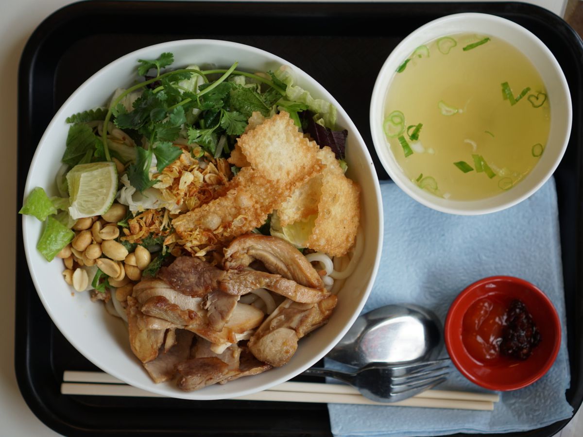 A dish of noodles, pork, chicken, and a mess of herbs, with a side of hot stock