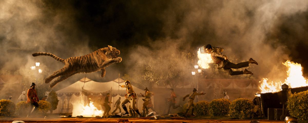A tiger and an Indian man with a crew cut, suspenders, a dress shirt holding a torch leap into the air at each other while soldiers cower behind them in RRR