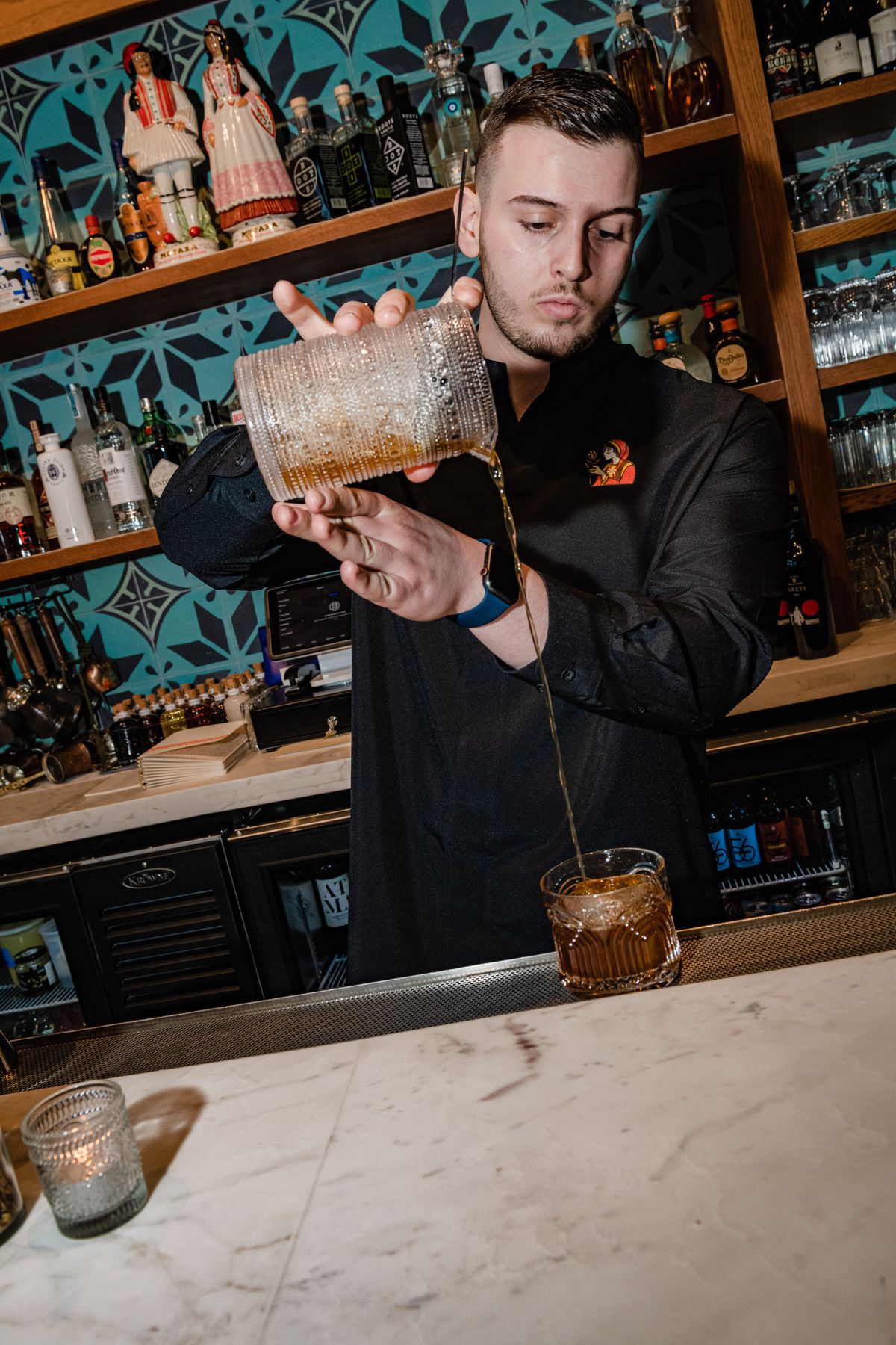 A man stands in a black uniform pouring a brown liquid into a cocktail glass.