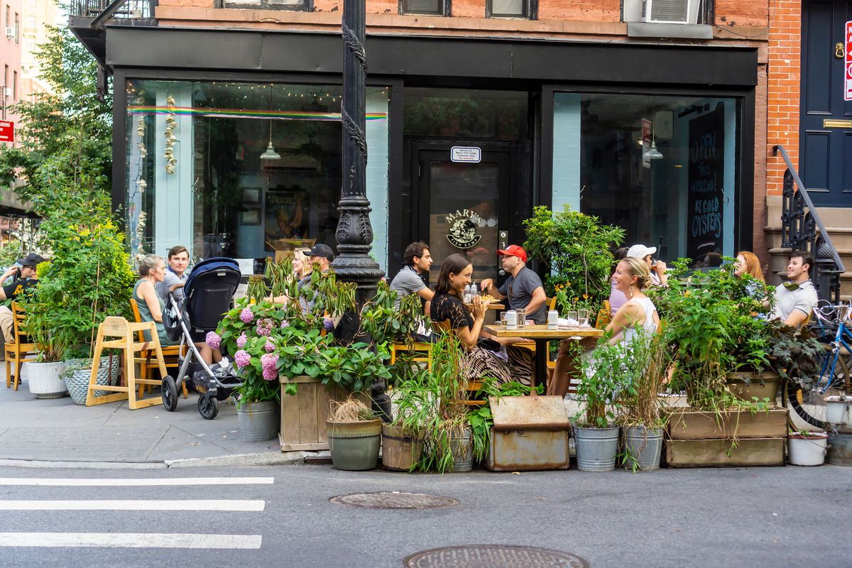 An outdoor dining set up on the street, featuring multiple tables of people surrounded by potted plants
