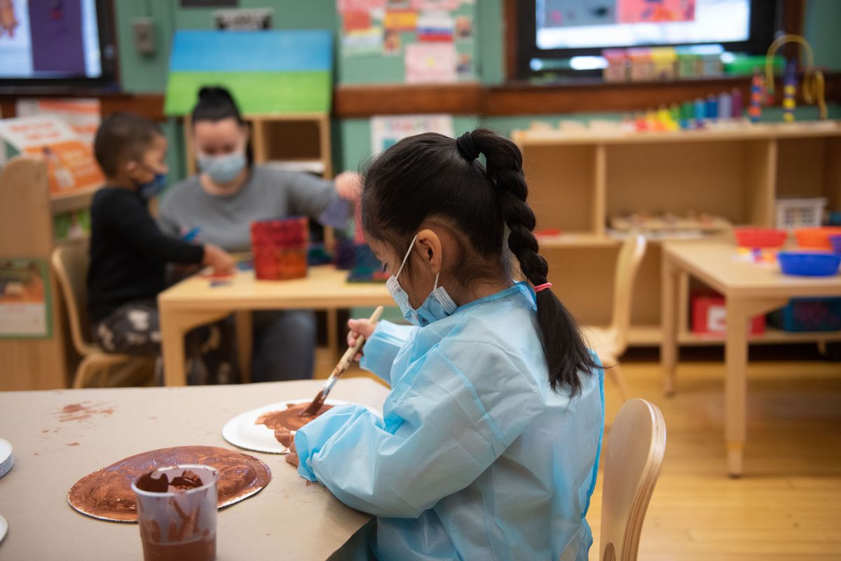 A young girl, wearing a blue smock and mask, paints at a wooden desk in a pre-school classroom