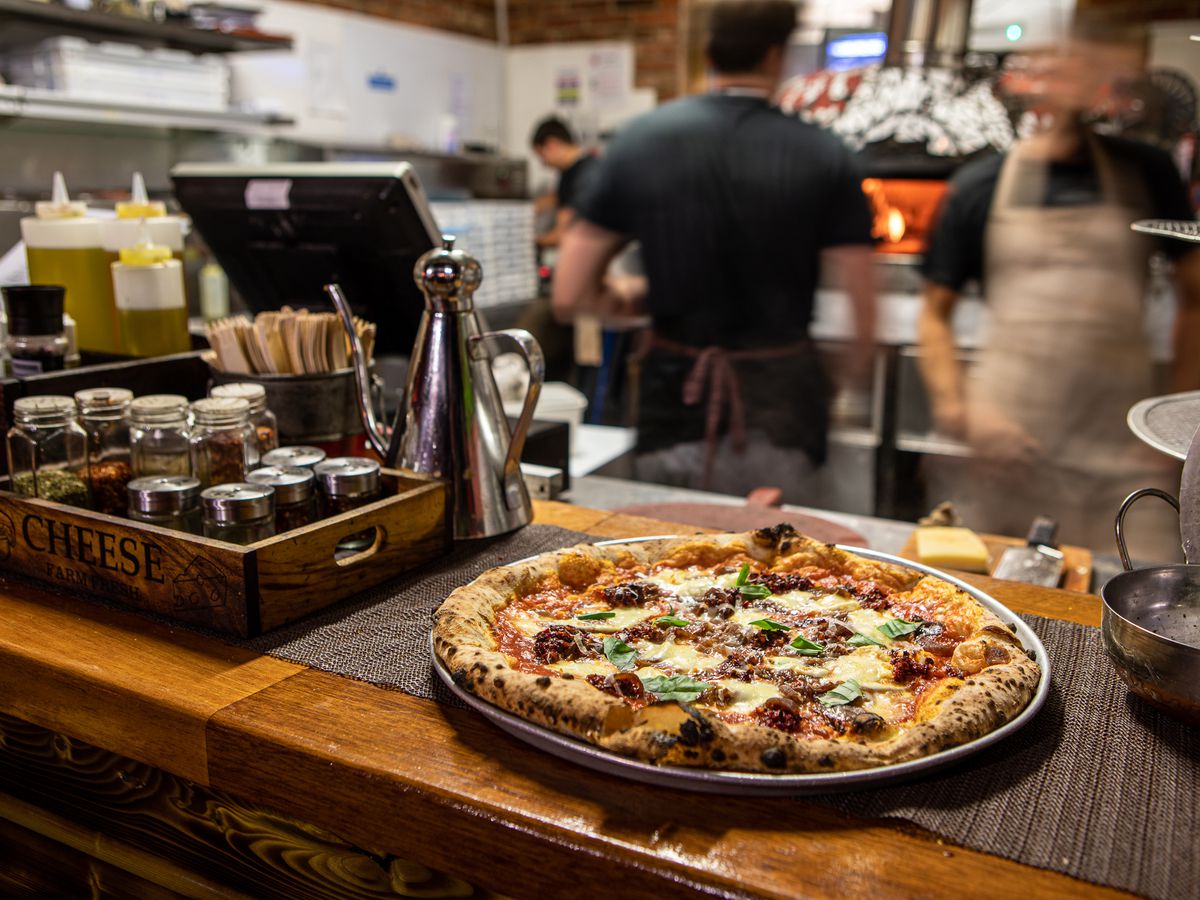 A full pizza lies on a serving dish on a wooden counter beside a tray of spice jars and an ornate olive oil dispenser, in front of a blurred out kitchen with staff at work