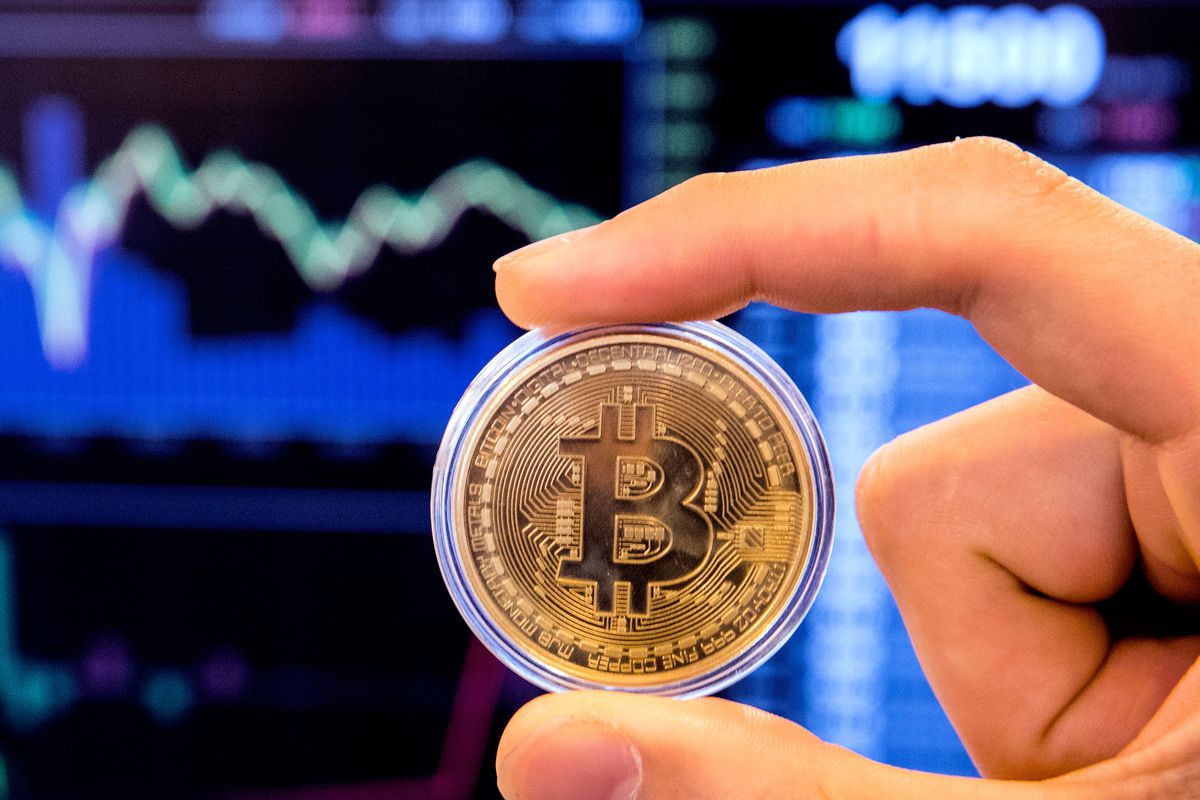 A gold-colored bitcoin coin held in a person’s finger and thumb, with stock market screens in the background