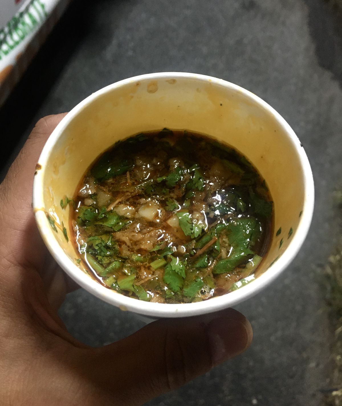 A hand holds a to-go cup of meaty consomme soup with cilantro, onion, and fatty spots swirling about