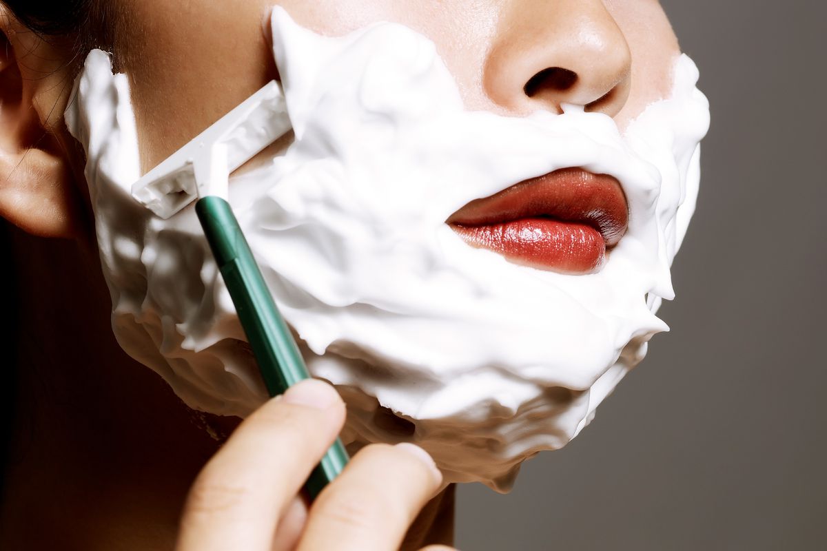 Lower half of a woman’s face is covered in shaving cream as she uses a razor to shave her face