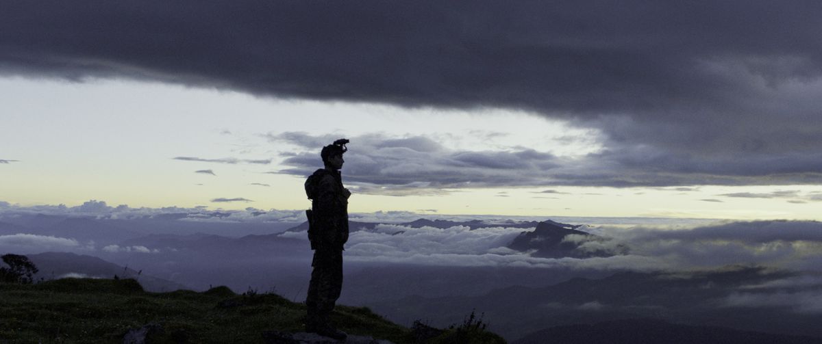 a boy stands atop a mountain in military gear, silhouetted by the setting sun