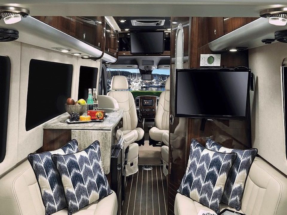 The interior of a RV camper van. There are two couches with pillows. There is a large television attached to one of the walls. There is a countertop with food. In the other room is the driving area.