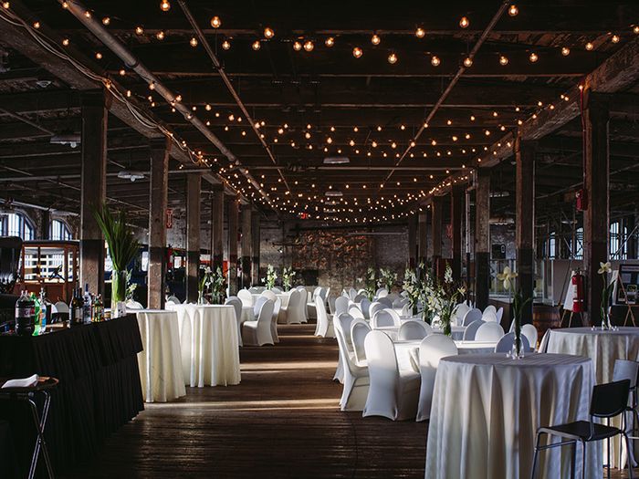 A large event space that is decorated for a party. There are many tables and chairs. Strings of holiday lights and strung across the ceiling above the tables and chairs.
