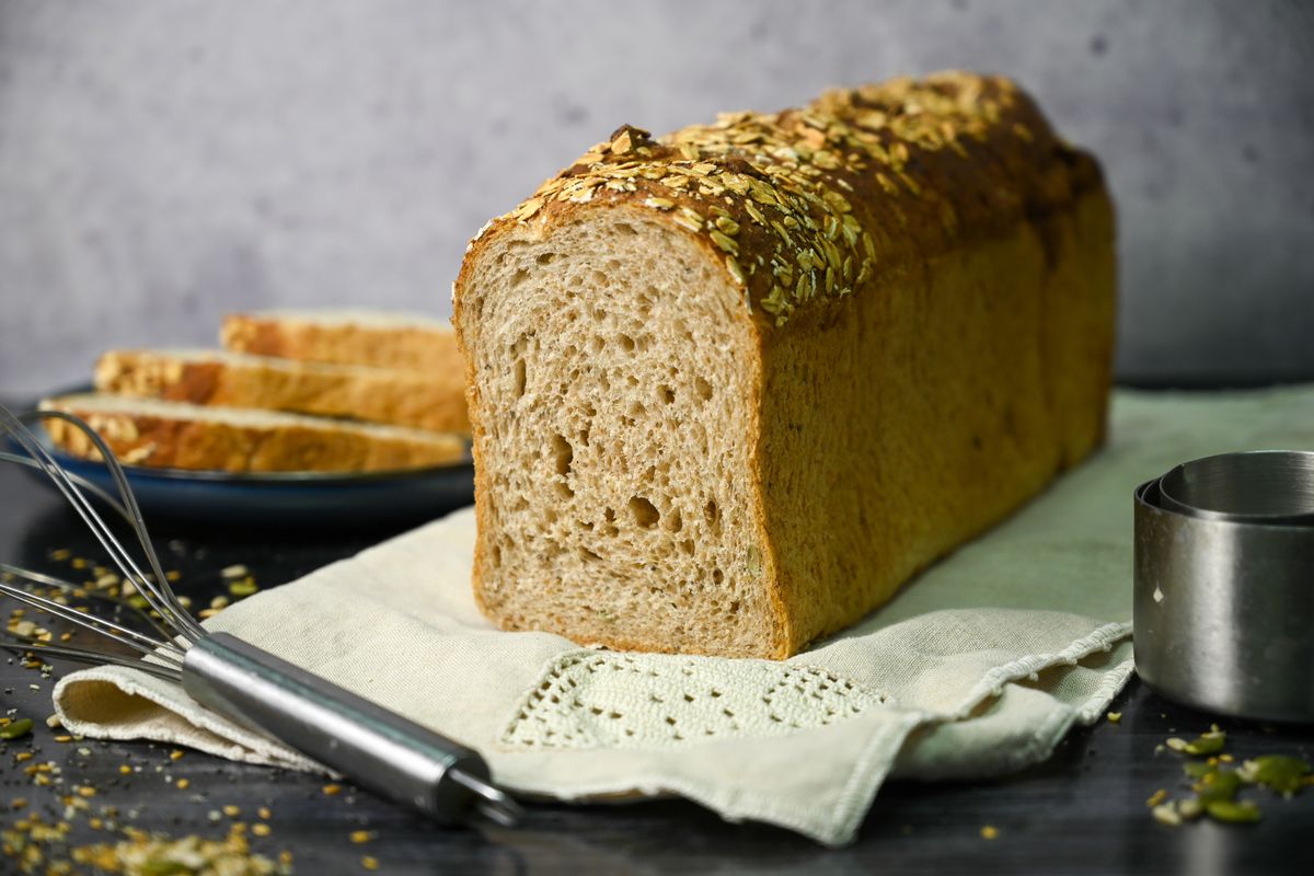 A tall, medium-brown loaf of bread is sliced near the end, with a stack of three slices piled up in the background. The loaf sits on an off-white cloth on a dark surface, with a metal whisk visible in the foreground.