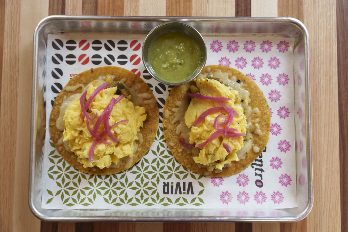 Two breakfast tacos made with eggs and garnished with purple onion sit in a silver tray on patterned paper. They have a side of salsa verde.