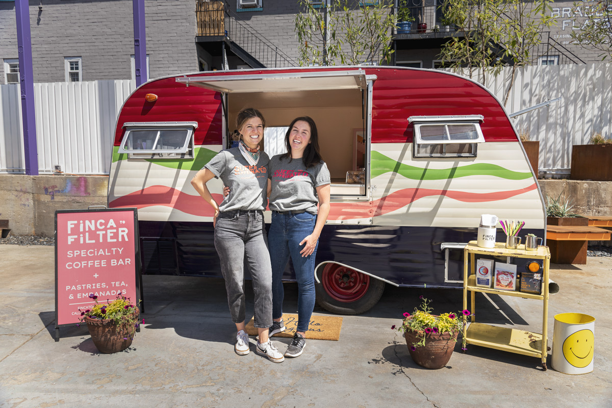 Two women stand in front of a colorfully decorated vintage trailer that has been repurposed into a coffee bar. 