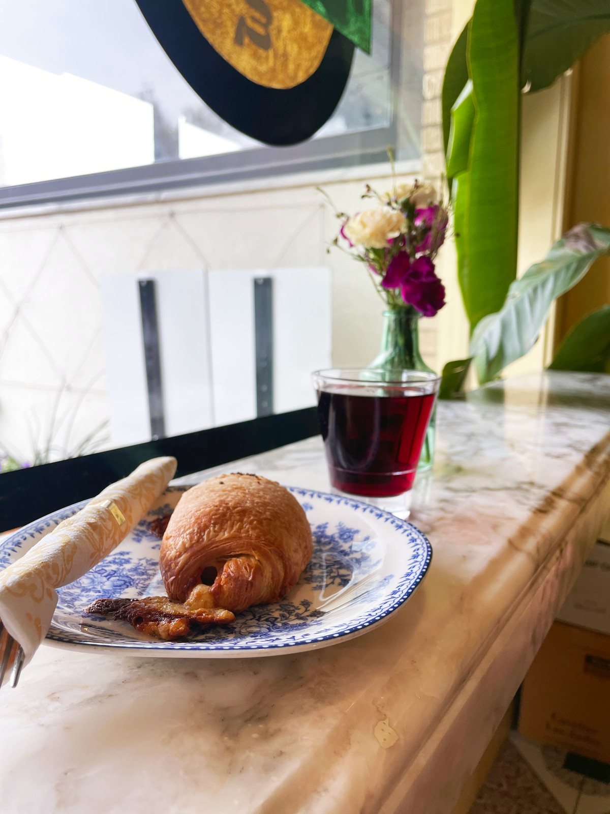 On a marble counter top, a blue and white plate hold a pastry, silverware in a napkin, a tumbler of red wine, and a vase of white and pink flowers.