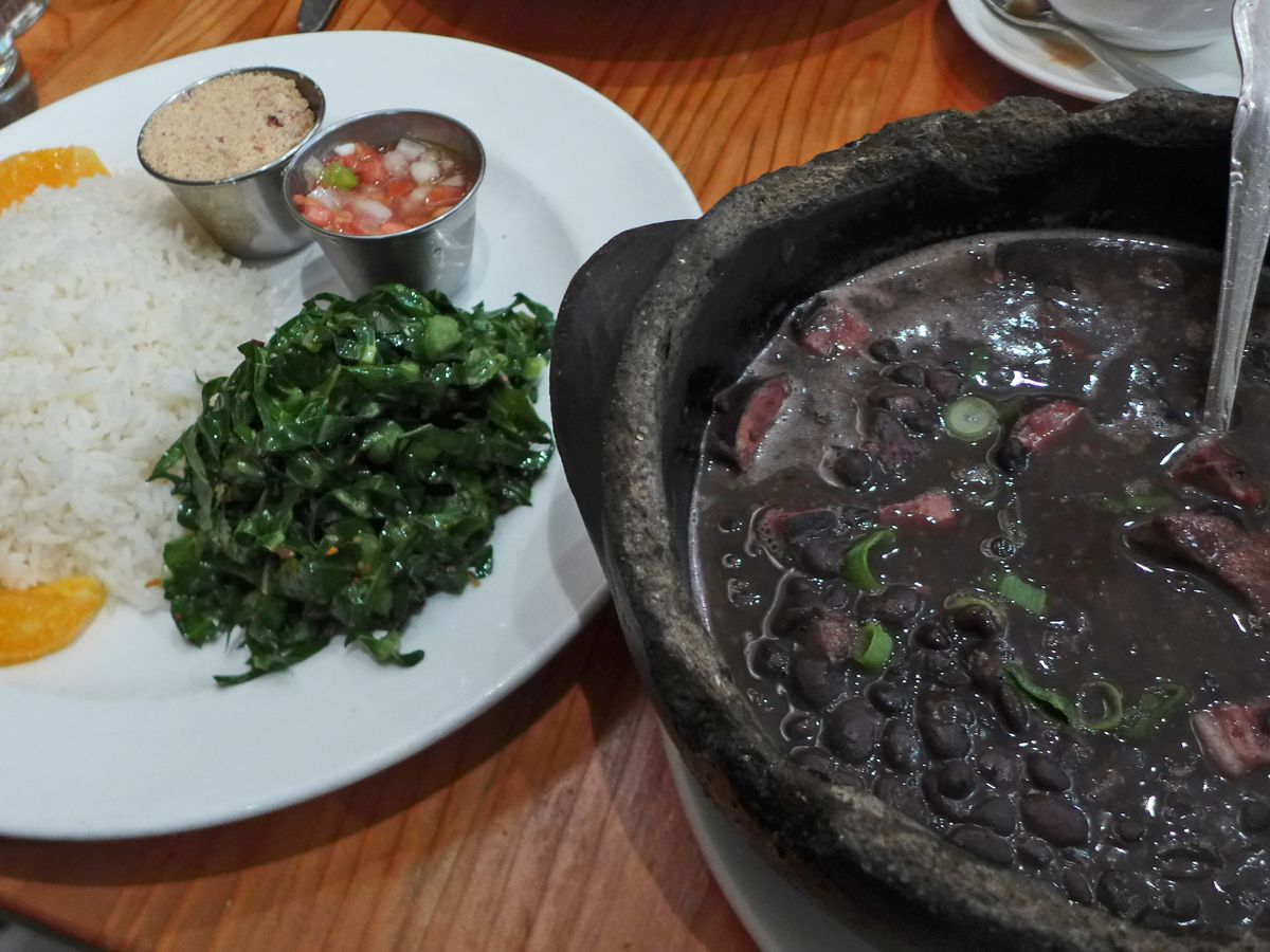 On the right a cast iron pot with pork and black bean stew, on the left a plate of rice, greens, and orange segments.