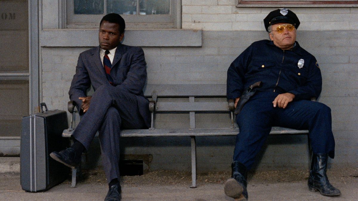 A man in a blue suit (Sidney Poitier) and a police officer (Rod Steiger) sit at opposite ends of a navy blue bench bolted to the floor against a painted brick wall.