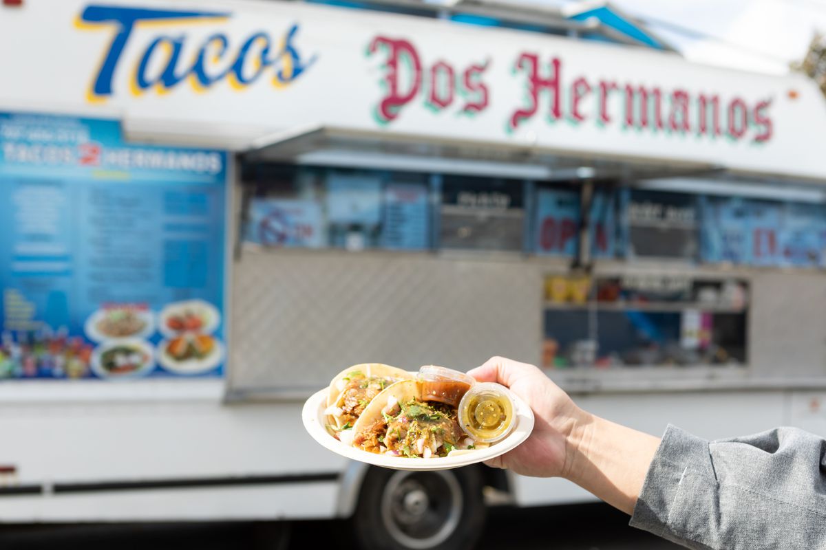A hand holding a paper plate of tacos in front of a truck.